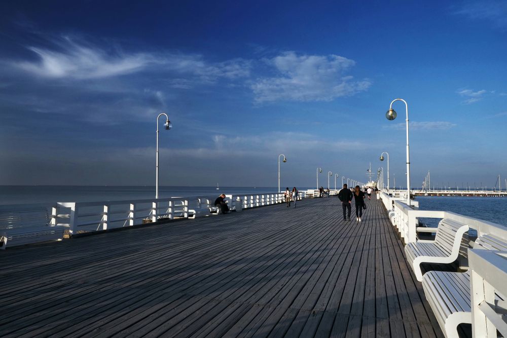 A zoomed out image of a beach promenade with people walking along it. The sky is blue with some white clouds and along the edge of the prom are white wooden benches.